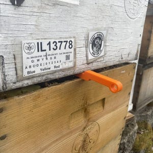 May include: A white wooden beehive with a label that reads "IL13778" and a sticker that says "Saint Ambrose Pray For Us". An orange beehive tool is placed on top of the hive.