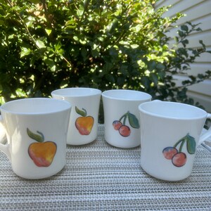 May include: Set of four white ceramic mugs with fruit designs. Two mugs feature a red and yellow cherry design, and two mugs feature a yellow and orange apple design.