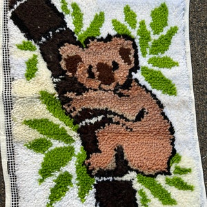 Könnte beinhalten: Ein brauner und rosa Koalabären-Teppich mit grünen Blättern, gestickt auf weißem Hintergrund. Der Teppich ist mit einem weißen Rand eingefasst.