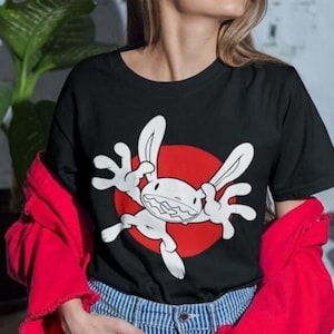 Può includere: Una maglietta nera con un disegno di coniglio bianco dei cartoni animati. Il coniglio indossa un cerchio rosso attorno al suo corpo. La maglietta è indossata da una persona con i capelli lunghi e castani.