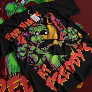 Peut inclure: T-shirt noir avec un graphisme coloré d'un personnage ressemblant à un ours et le texte "FIVE NIGHTS AT FREDDY'S". Le design comprend des accents verts, jaunes, rouges et violets. Une étiquette blanche indique une taille L.