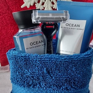 以下が含まれることがあります： 青いタオル、バス＆ボディワークスのオーシャンボディウォッシュボトル、バス＆ボディワークスのオーシャンボディローションチューブ、そしてカミソリが入った青と白のギフトバスケット。バスケットは白い雪の結晶で飾られています。
