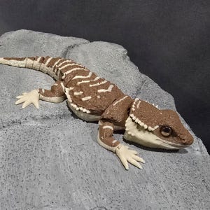 Peut inclure: Une figurine de lézard articulée de couleur brune et crème avec un corps texturé et des yeux sombres, posée sur une structure grise à la surface rugueuse imitant la roche. Le lézard présente des articulations segmentées sur le dos et la queue.