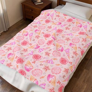 Puede incluir: Una manta suave con un patrón repetitivo de conchas marinas, estrellas de mar y peces pequeños estilo acuarela en tonos rosa, coral y amarillo sobre un fondo rosa claro, extendida sobre una cama con una almohada blanca y una mesita de noche.