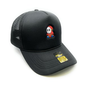 May include: Black trucker hat with a red and blue character embroidered on the front. The character is wearing a mask and has a red and white hat. The hat has a black mesh back and a black brim. The hat has a yellow tag on the brim that says "Official Licensed Product" and "Hatzone".