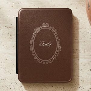 May include: A dark brown rectangular e-reader case with a textured surface. The center features an engraved oval frame with decorative scrollwork details and the name Emily written in a cursive script font in the middle of the oval.