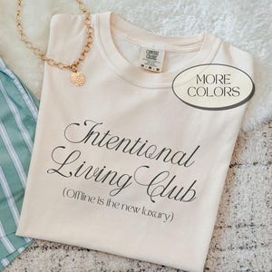 May include: Cream-colored t-shirt with the text "Intentional Living Club" and the tagline "(Offline is the new luxury)". A gold necklace with a pendant and green striped shorts are also visible.