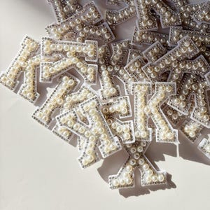 May include: A collection of letter "K" patches, each adorned with faux pearls and silver-colored rhinestones. The patches are arranged on a white surface, creating a sparkling and elegant display. These patches are ideal for adding a personalized touch to clothing or accessories.