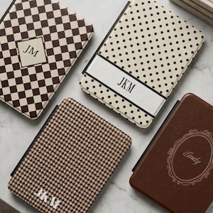 May include: Four rectangular e-reader cases with distinct patterns: one brown and white checkerboard with JM, one cream with black polka dots and JKM, one brown and white houndstooth with JKM, and one solid brown with an oval frame and Emily.