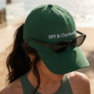 May include: A forest green baseball cap featuring the white embroidered text SPF & Chardonnay on the front crown. A pair of black sunglasses with dark lenses is resting horizontally across the front of the cap, positioned above the brim.