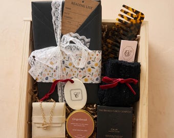 May include: A wooden gift box containing a black wrapped item with a floral band and white lace ribbon, a brown reading list card, a tortoise shell comb, a small pink box, black fuzzy socks with a red ribbon, a gold chain, a gingerbread candle, and soap.