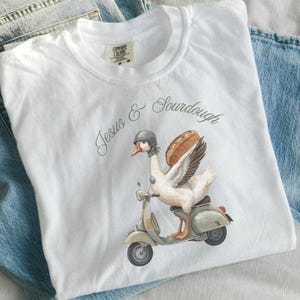 May include: White t-shirt with a whimsical illustration of a goose wearing a helmet and riding a scooter, with a loaf of bread as wings. The text "Jesus & Sourdough" is above the graphic. The shirt is laid on top of blue jeans.