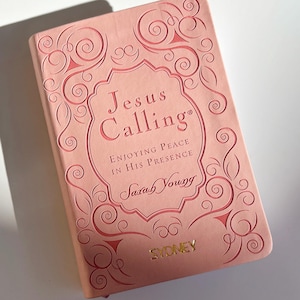 May include: A pink book titled "Jesus Calling" with the subtitle "Enjoying Peace in His Presence" by Sarah Young. The cover features an ornate design and the word "SYDNEY" in gold lettering.
