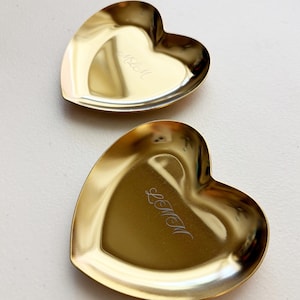 May include: Two heart-shaped gold-toned metal trinket dishes with a polished finish. Each dish features a delicate, cursive script engraving in the center. The dishes have smooth, curved edges and a reflective surface that catches the light.