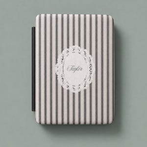 May include: A rectangular e-reader case featuring a vertical black and white pinstripe pattern. Centered on the front is a white oval lace-style emblem with the name Taylor printed in a cursive script font. A black spine is visible on the left.
