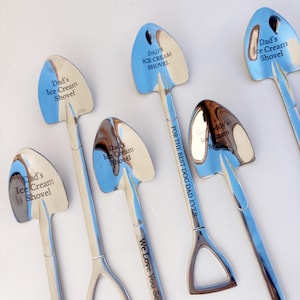 May include: A collection of silver-toned metal spoons shaped like miniature shovels. The bowls are engraved with the text Dad's Ice Cream Shovel, while one handle features the text FOR THE BEST DOG DAD EVER and We Love You Scoopfuls!