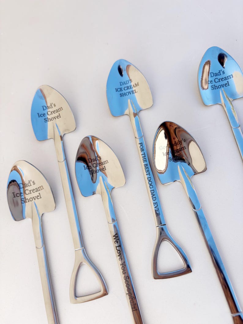 May include: A collection of silver-toned metal spoons shaped like miniature shovels. The bowls are engraved with the text Dad's Ice Cream Shovel, while one handle features the text FOR THE BEST DOG DAD EVER and We Love You Scoopfuls!