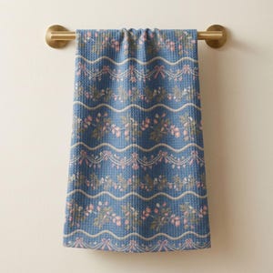 May include: A blue waffle-weave hand towel hanging on a brass-toned metal bar. The towel features a repeating pattern of horizontal wavy cream-colored lines, pink bows, and clusters of small pink and green floral sprigs across the fabric.