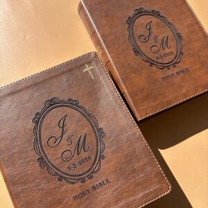 May include: Two brown leather Holy Bibles with gold crosses and decorative oval designs. The oval design contains the initials "J & M" and the date "5-2-2026". The words "Holy Bible" are printed at the bottom of the cover.