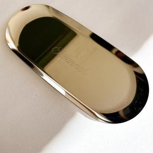 May include: An oval-shaped gold-colored metal tray with a polished, reflective surface. The name Savannah is engraved in a cursive script font in the center of the tray. The tray features raised edges and a smooth, metallic finish throughout.