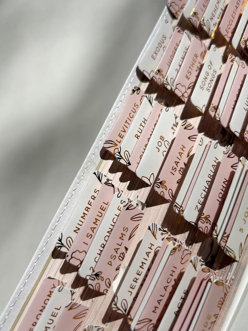 K&ouml;nnte beinhalten: Nahaufnahme von Bibel-Reitern mit goldfarbenen Buchstaben und floralen Designs. Die Reiter sind in Rosa- und Wei&szlig;t&ouml;nen gehalten und tragen Titel wie "Levitikus", "Ruth" und "Hiob". Die Reiter sind f&auml;cherf&ouml;rmig angeordnet, was auf ein Buch hindeutet.
