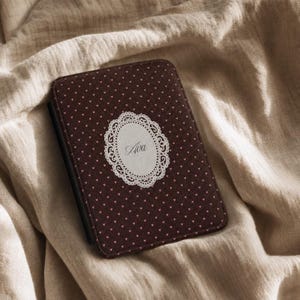 Pode incluir: Uma capa retangular para e-reader em castanho escuro com um padrão repetitivo de pequenos corações cor-de-rosa claro. Ao centro, uma etiqueta oval branca com uma borda decorativa recortada exibe o nome Ava em letra cursiva preta.