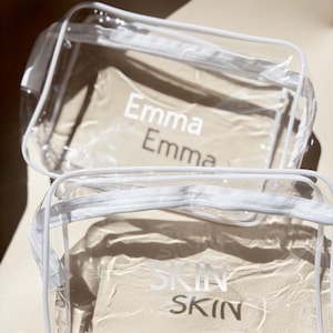 May include: Two clear, rectangular cosmetic bags with white trim and zippers. The top bag has the name "Emma" printed twice in white. The bottom bag has the word "SKIN" printed twice in white. Both bags are filled with a beige material.