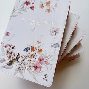 K&ouml;nnte beinhalten: Eine wei&szlig;e Bibel mit floralen Illustrationen in Rosa, Orange und Blau. Auf dem Einband steht "THE SPIRITUAL GROWTH BIBLE" in goldener Schrift. Die Seitenr&auml;nder sind rosa und gold.