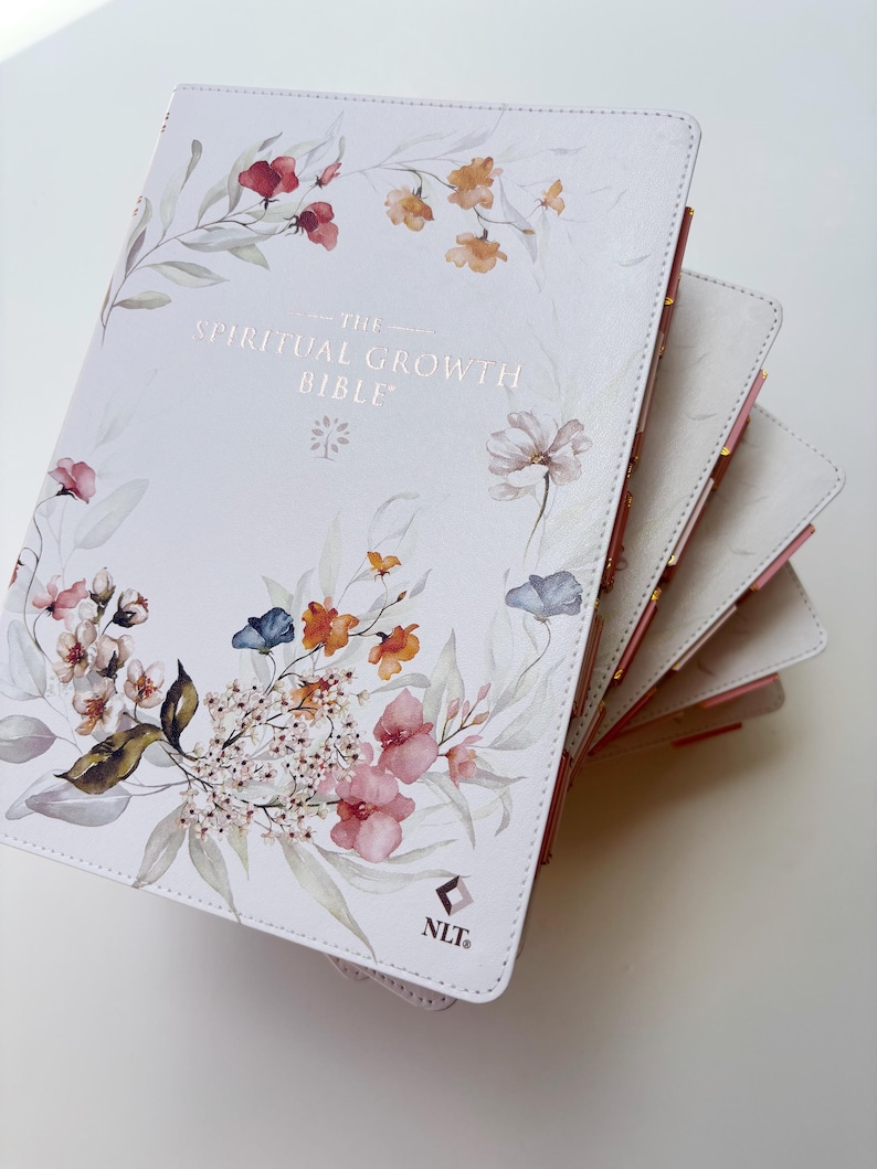 K&ouml;nnte beinhalten: Eine wei&szlig;e Bibel mit floralen Illustrationen in Rosa, Orange und Blau. Auf dem Einband steht "THE SPIRITUAL GROWTH BIBLE" in goldener Schrift. Die Seitenr&auml;nder sind rosa und gold.
