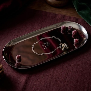 May include: An oval-shaped silver-toned metal tray with a polished reflective surface. The center features an engraved decorative frame containing the letter P. Small red berries and a miniature pinecone are placed on the tray surface.
