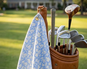 Op de afbeelding: Een lichtblauwe golfhanddoek met een wit bloemen- en golfkarretjespatroon, voorzien van een goudkleurige metalen oogje en ring, hangend aan een bruine leren golf-tas gevuld met diverse metalen golfclubs en een houten driver.