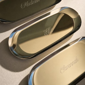 May include: Three oval-shaped gold-toned metal trays with polished reflective surfaces. Each tray features a unique name engraved in a cursive script: Madison, Elle, and Savannah. The trays have raised edges and a smooth, metallic finish.