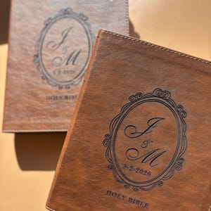 May include: Two brown leather Holy Bibles. The Bibles have a decorative oval design with the initials "J & M" and the date "5-2-2026" engraved on the cover. One Bible has a cross on the cover.