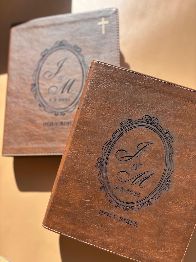May include: Two brown leather Holy Bibles. The Bibles have a decorative oval design with the initials "J & M" and the date "5-2-2026" engraved on the cover. One Bible has a cross on the cover.