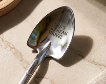 Op de afbeelding: Een zilverkleurige metalen lepel in de vorm van een kleine tuinschep. De kom van de lepel is voorzien van de gegraveerde tekst DAD'S ICE CREAM SHOVEL in hoofdletters. Het bestek heeft een gladde, gepolijste afwerking en een puntige tip.