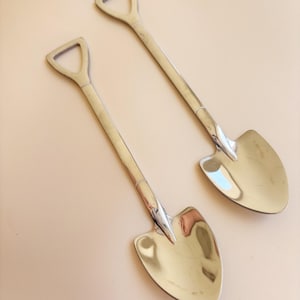 May include: Two silver-colored metal spoons with handles shaped like the shafts of shovels and bowls shaped like spade blades. The spoons have a polished, reflective finish and a distinct D-shaped handle top, mimicking the design of a garden tool.