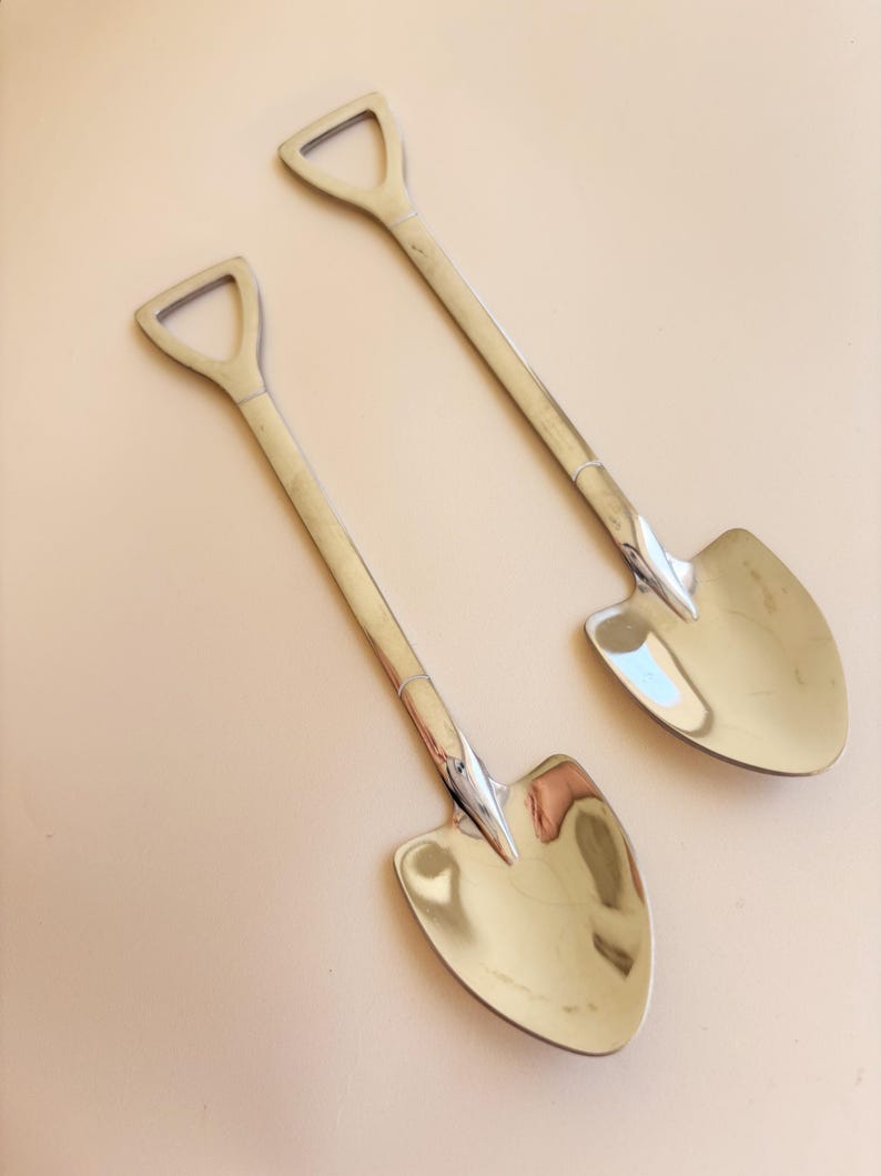 May include: Two silver-colored metal spoons with handles shaped like the shafts of shovels and bowls shaped like spade blades. The spoons have a polished, reflective finish and a distinct D-shaped handle top, mimicking the design of a garden tool.
