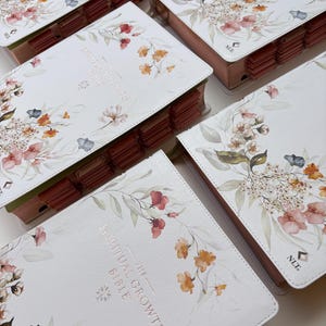 May include: Several copies of "The Spiritual Growth Bible" are displayed. The white covers feature floral watercolor designs in shades of pink, orange, and blue. The book's edges are a soft pink, and the pages have labeled tabs.