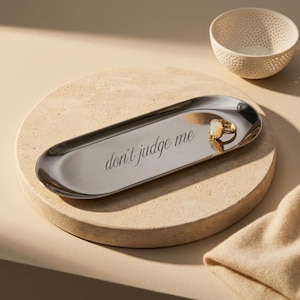 May include: An elongated, oval-shaped silver-toned metal tray featuring the engraved text "don't judge me" in a cursive script. A gold-toned ring with a rectangular stone rests on the surface of the tray, which has a smooth, reflective finish.