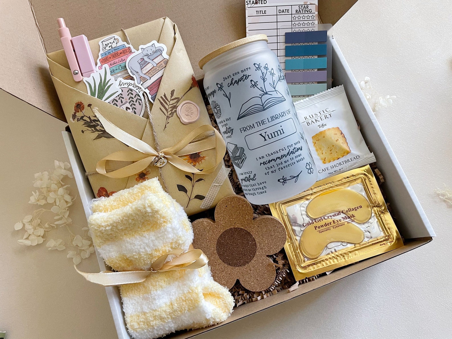 Box with a variety of gifts tied in yellow ribbon. A personalized cup with bookish icons, eye masks, stucky notes for annotating, a pair of socks, and a flower coaster, and brown paper wrapped around a book folded in sections on the front holding stickers a pet, and a highlighter are all in the box with paper confetti.