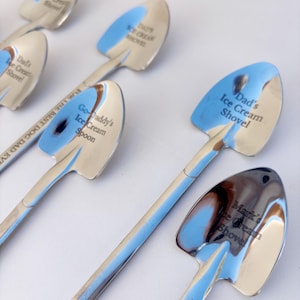 May include: A collection of silver-toned metal spoons shaped like miniature shovels. Each spoon features custom engravings including Dad's Ice Cream Shovel, Go-Daddy's Ice Cream Spoon, and Mark's Ice Cream Shovel, with one spoon engraved with FOR THE BEST DOG DAD EVER.