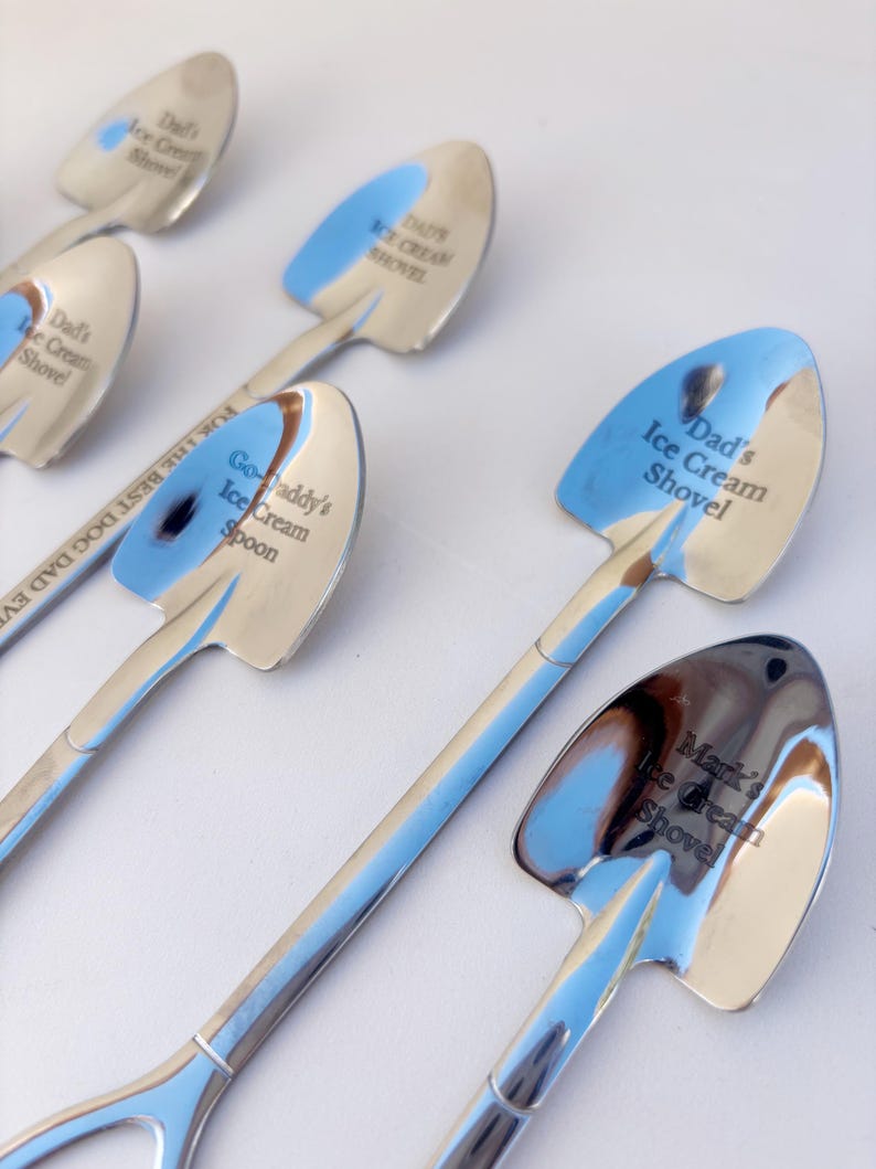 May include: A collection of silver-toned metal spoons shaped like miniature shovels. Each spoon features custom engravings including Dad's Ice Cream Shovel, Go-Daddy's Ice Cream Spoon, and Mark's Ice Cream Shovel, with one spoon engraved with FOR THE BEST DOG DAD EVER.