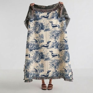 May include: A woven throw blanket featuring a blue and cream toile pattern with repeating illustrations of dachshund dogs, trees, and gazebo structures. The rectangular blanket has a decorative fringed edge along the top and bottom borders.