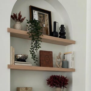 May include: A built-in arched shelf unit with three wooden shelves. The shelves display various decorative items, including vases with dried flowers, a framed picture, books, a decorative bowl, a statue, and woven boxes. The color scheme is neutral with pops of burgundy and black.
