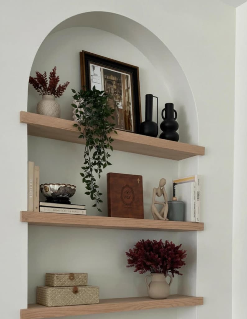 May include: A built-in arched shelf unit with three wooden shelves. The shelves display various decorative items, including vases with dried flowers, a framed picture, books, a decorative bowl, a statue, and woven boxes. The color scheme is neutral with pops of burgundy and black.
