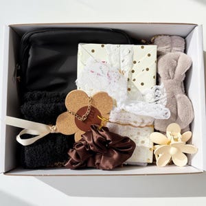 May include: A white gift box filled with various items. Includes a black pouch, black socks with a white ribbon, a wrapped gift with lace, a brown scrunchie, a flower-shaped hair clip, and a plush bunny ear headband.