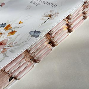 K&ouml;nnte beinhalten: Eine wei&szlig;e Bibel mit floralen Illustrationen und dem Text "The Spiritual Growth Bible". Das Buch hat rosa und goldfarbene Seiten mit Kapitelreitern. Das Cover zeigt Blumen im Aquarellstil in Orange-, Rosa- und Blaut&ouml;nen.