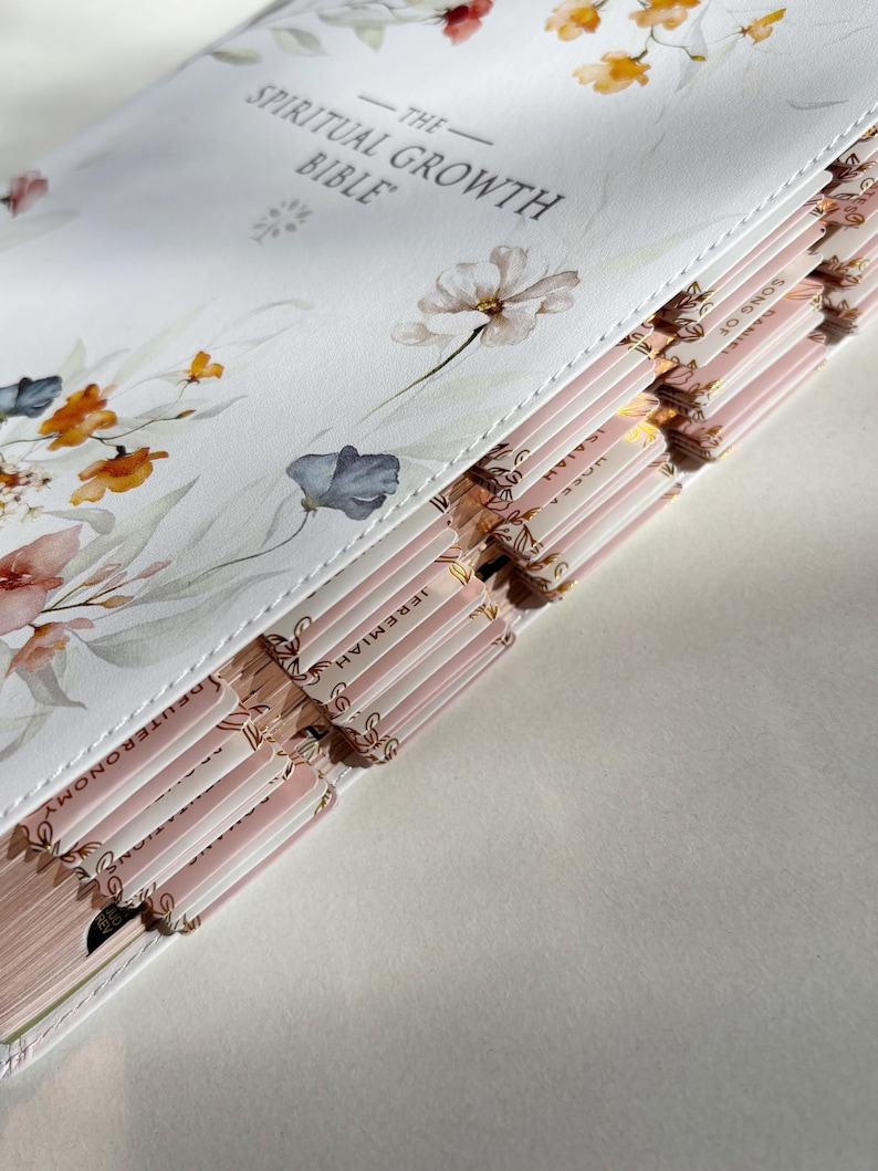 K&ouml;nnte beinhalten: Eine wei&szlig;e Bibel mit floralen Illustrationen und dem Text "The Spiritual Growth Bible". Das Buch hat rosa und goldfarbene Seiten mit Kapitelreitern. Das Cover zeigt Blumen im Aquarellstil in Orange-, Rosa- und Blaut&ouml;nen.