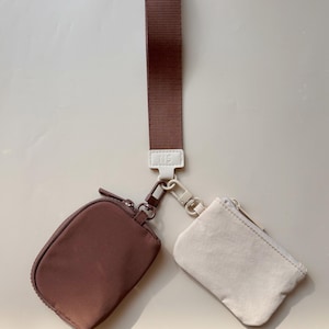 May include: A brown wrist strap with a white tag and two small zippered pouches. One pouch is brown, and the other is off-white. The pouches are connected to the strap with metal clasps.