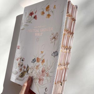 K&ouml;nnte beinhalten: Eine wei&szlig;e Bibel mit floralen Aquarellillustrationen und dem Text "THE SPIRITUAL GROWTH BIBLE". Das Buch hat rosafarbene Seiten und Register. Der Einband zeigt Blumen in Rot, Orange und Blau.